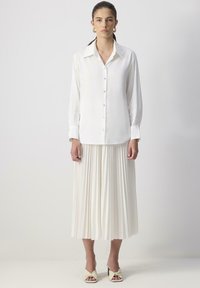 Femeie îmbrăcată cu o bluză albă cu mâneci lungi și nasturi, fustă midi pliată, cercei rotunzi aurii și sandale cu toc crem, stând pe un fundal simplu.