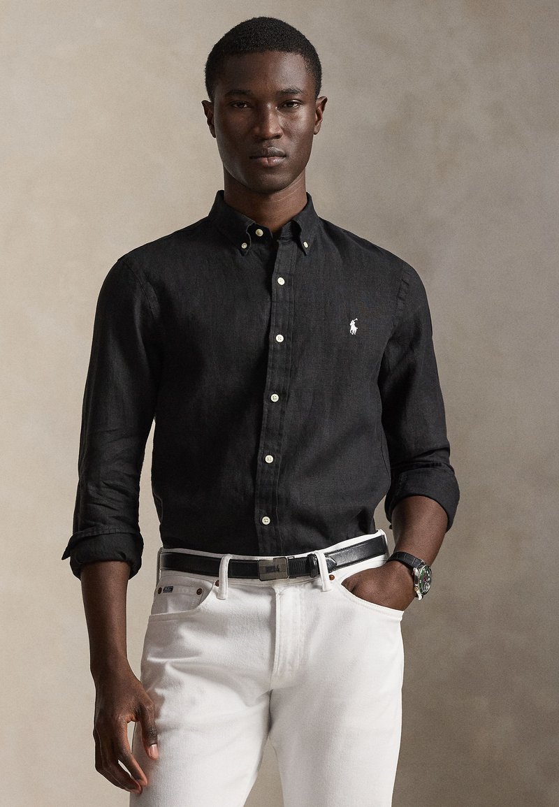 Άνδρας που φοράει μαύρο πουκάμισο  με κουμπιά και μακριά μανίκια, γυρισμένα μανίκια, λευκό παντελόνι, μαύρη ζώνη και ρολόι χειρός, στέκεται μπροστά σε ουδέτερο φόντο.