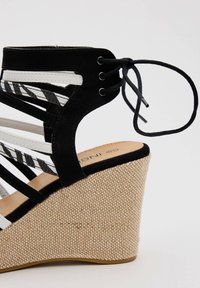 Sandalia cuña en blanco y negro con una plataforma texturizada similar a la paja, correas de ante y detalle de cordones en el tobillo.