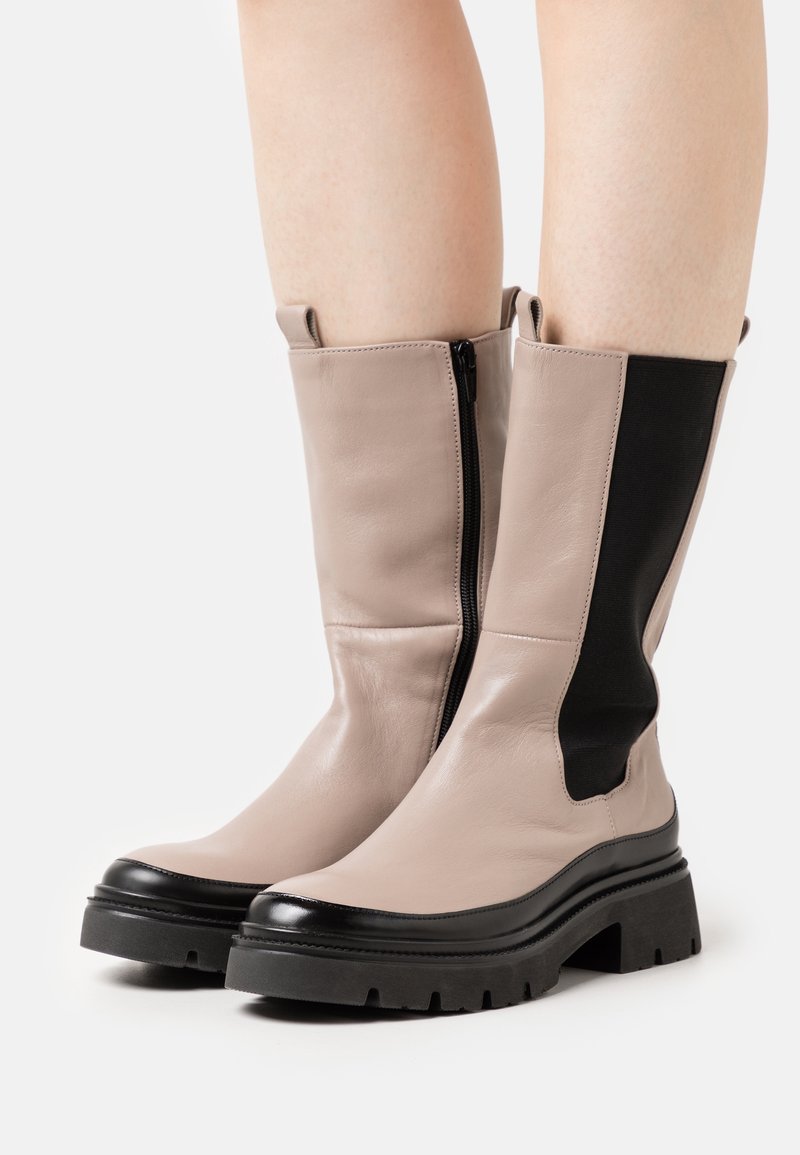 Knee-high boots made of light beige leather with black elastic side panels, featuring a chunky black rubber sole and side zipper detail.