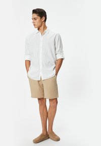 Camicia bianca in lino con maniche arrotolate, pantaloni beige texturizzati e scarpe slip-on color camel, in piedi su uno sfondo semplice.