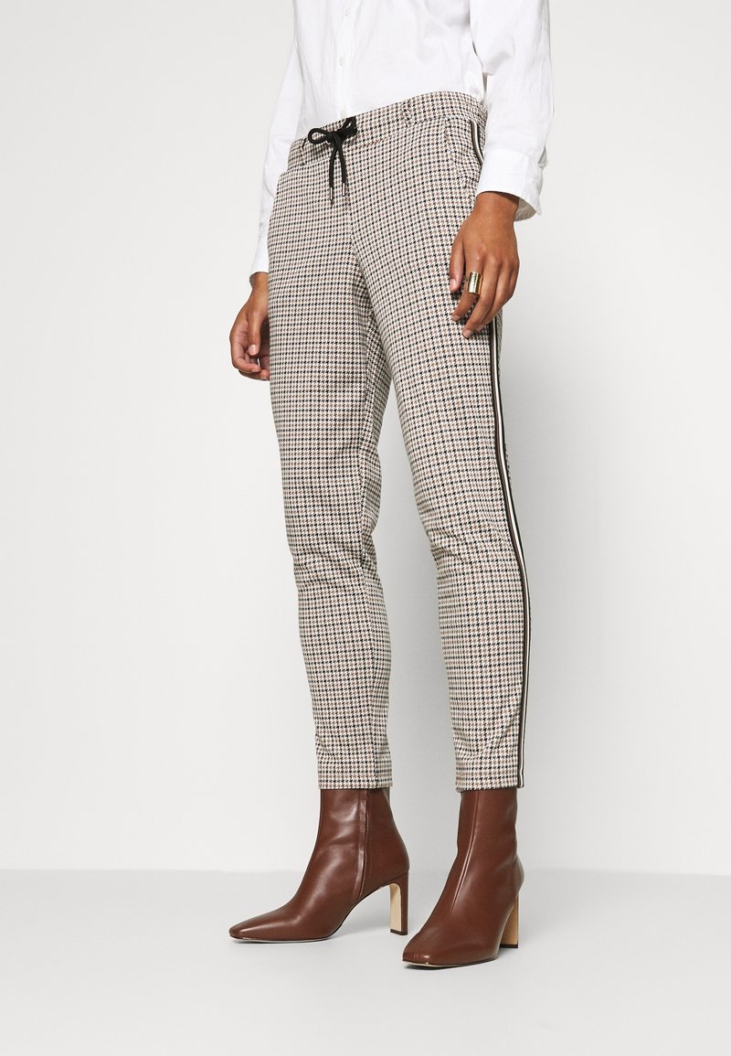 Femme portant un pantalon slim beige à motif pied-de-poule avec des bandes latérales noires et des bottines en cuir marron à talons, associé à une chemise blanche.