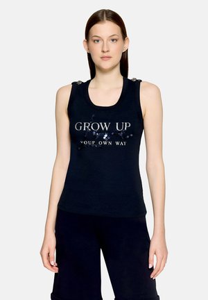 Kobieta w czarnym topie bez rękawów z srebrnym napisem „GROW UP YOUR OWN WAY” i czarnych szortach, stojąca na jasnym, jednolitym tle.