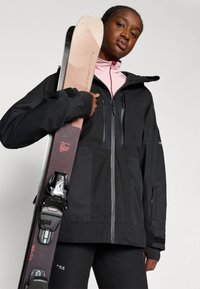 Casaco de esqui preto à prova d'água com zíperes cinzentos, combinado com camadas internas rosas. Um par de skies com fixações pretas exibido de forma proeminente.