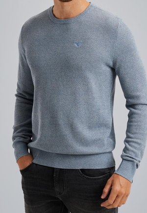 Homme portant un pull en maille texturée bleu clair avec poignets et ourlet côtelés, associé à un jean noir, debout les mains près des poches.