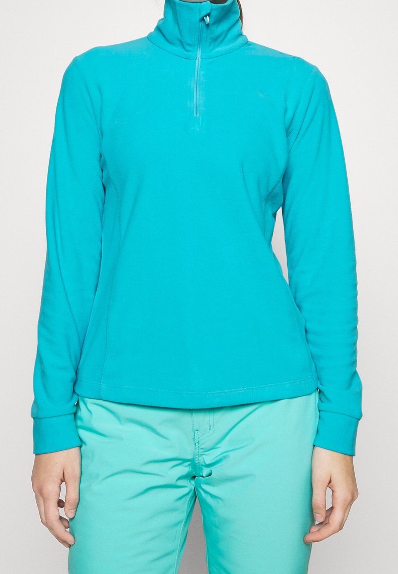 Femme portant un haut polaire turquoise à manches longues avec fermeture éclair quart, et un pantalon bleu clair, debout devant un fond uni clair.