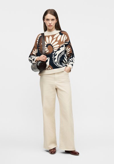 Γυναίκα που φοράει πουλόβερ με σχέδιο σε καφέ, εκρού και ναυτικό μπλε χρώμα, φαρδιά παντελόνια εκρού, καφέ παπούτσια και κρατάει μια στρογγυλή σκουρόχρωμη καφέ τσάντα στον ώμο της.