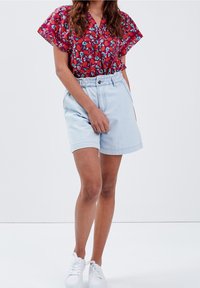 Donna con una maglietta rossa a fiori a maniche corte, pantaloni corti a vita alta blu chiaro e sneakers bianche, in piedi contro uno sfondo chiaro e semplice.