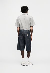 Άνδρας με λεύκη που φορά ριγέ κοντομάνικο πουκάμισο, σκούρα τζιν σορτς μέχρι το γόνατο, λευκές κάλτσες και μπεζ αθλητικά παπούτσια, γυρισμένος με την πλάτη σε απλό φόντο.