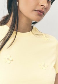 Gelbes T-Shirt mit rundem Halsausschnitt, das auf der Vorderseite drei Applikationen aus Stoffblüten mit klaren Verzierungen aufweist. Glatte Textur.