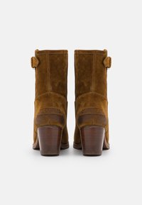 Botas de tobillo de ante marrón con un tacón ancho, que presentan una superficie lisa, detalles laterales y un logotipo en la parte trasera.