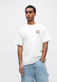 Λευκή βαμβακερή μπλούζα με στρογγυλή λαιμόκοψη, κοντά μανίκια και τυπωμένο σχέδιο στο αριστερό στήθος. Φορεμένη με ανοιχτό μπλε τζιν σορτς.