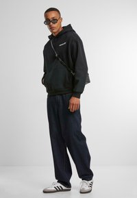 Svart hettegenser med hvitt logo, parret med løse mørke denimjeans. Tilbehøret inkluderer svart skulderveske og grå sneakers med hvite striper.