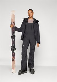 Schwarze Skijacke und Latzhose mit mehreren Taschen, getragen mit schwarzen Skischuhen. Person hält Skier mit Bindungen. Neutrale Hintergrund.
