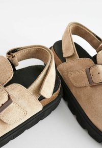 Sandales en suède beige avec brides ajustables, semelles en caoutchouc noir et finition texturée. Comprend un accent de boucle et une semelle intérieure rembourrée.