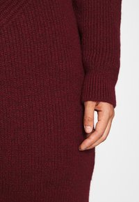 Pull en maille bordeaux avec une texture côtelée et des manches ajustées. Main placée sur la taille, mettant en valeur le design ajusté du vêtement.