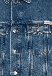 Jeans jakna v srednji modri barvi, ki ima srebrno-tonirane kovinske gumbe, dvojna žepa na prsih in oranžne šive za kontrast. Prisotna oznaka Calvin Klein.