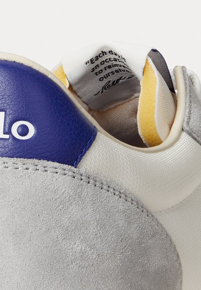 Close-up of sneaker heel showing gray suede, blue leather patch with partial white text, and fabric label with printed quote inside collar.
