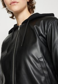 Giubbotto bomber in pelle nera con cappuccio, dotato di chiusura con zip, tasche laterali e una texture liscia. Decorato con lacci neri.