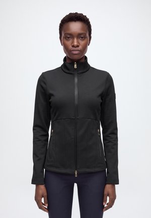 Une femme porte une veste noire zippée à col montant et poches zippées, devant un fond blanc uni.