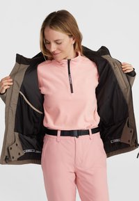 Bruine buitenjas met zwarte voering, lichtroze fleece top, roze broek en een zwarte riem. Rits en zakken zichtbaar in de jas.