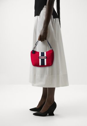 Femme portant des talons noirs et une jupe blanche, tenant un petit sac à main rouge avec des rayures verticales bleu marine et blanc et un logo au centre.