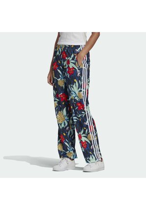 Person iført marineblå træningsbukser med rød og gul blomsterprint og hvide sidelister, kombineret med hvide sneakers og en hvid top.