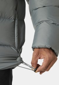 Grijze pufferjack met een doorgestikte textuur. Een hand trekt aan de koord bij de zoom, die een strakker mechanisme heeft. Glad stoffen afwerking.