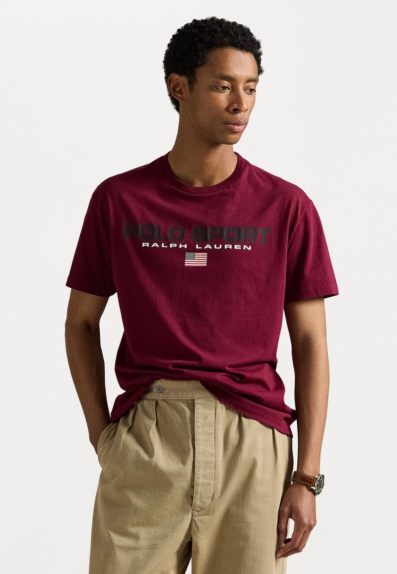 Bordová bavlnená tričko s grafikou "POLO SPORT" a "RALPH LAUREN"; krátke rukávy; voľný strih. V kombinácii s béžovými nohavicami.