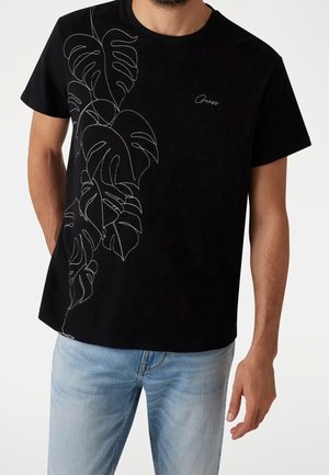 T-shirt en coton noir avec un contour blanc de feuilles de monstera sur le côté gauche. Manches courtes, coupe décontractée, col rond.