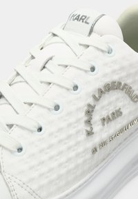 Hvit skinnsneaker med teksturerte paneler, flate skolisser og metallisk merke. Inkluderer lyseblå øyeluker og en polstret såle.