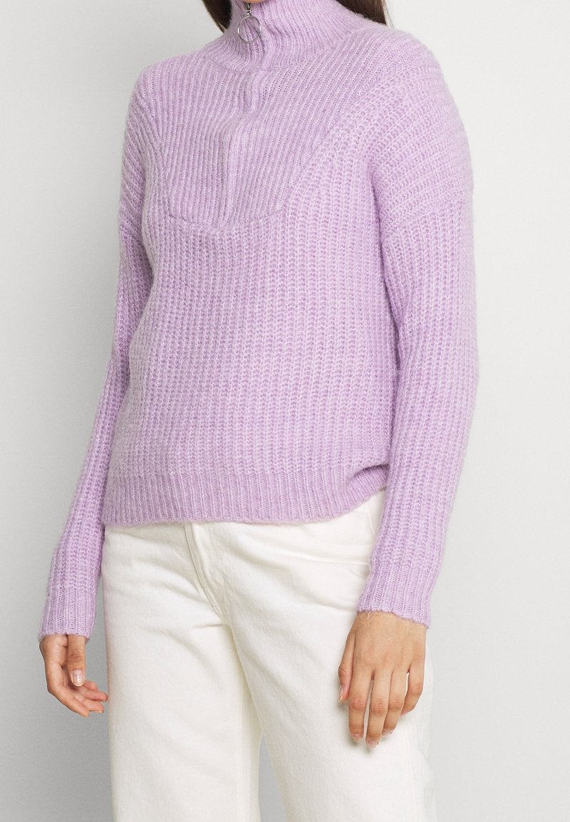 Femme portant un pull à col roulé en maille épaisse de couleur violet clair et un pantalon blanc cassé, debout devant un fond blanc uni.