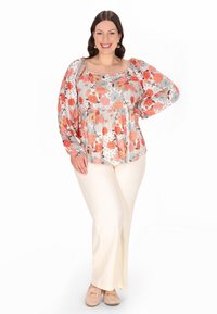 Blomstret bluse med puffede ærmer, der har en blanding af orange, blå og hvide mønstre, parret med krèmefarvede bukser og beige sko.