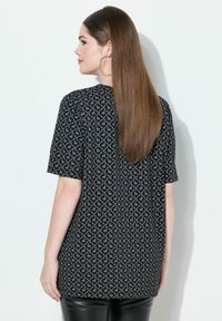 Černý top s krátkým rukávem a geometrickým bílým vzorem. Tkanina se zdá být měkká a strukturovaná, nabízí volný střih a zakřivený spodní lem.