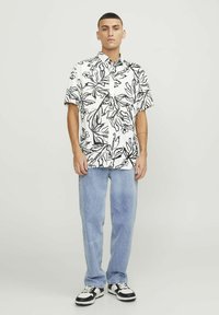 Chemise à manches courtes, boutonnée, blanche avec un motif de feuilles noires ; associée à un jean bleu clair ample et des baskets noir et blanc.