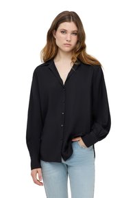 Camicia nera a maniche lunghe con bottoni in tessuto leggero, caratterizzata da un colletto a punta, una placca con bottoni frontale e una vestibilità rilassata. Abbinata a jeans blu chiaro.