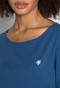 T-shirt in cotone blu con scollatura rotonda, caratterizzato da un piccolo logo bianco sul lato sinistro del petto. Texture liscia con dettagli di cucitura visibili.