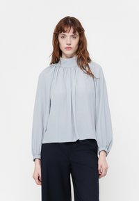 Hellgraue Bluse mit Schalkragen, gefältelter Vorderseite und gerafften langen Ärmeln. Der Stoff wirkt glatt und strukturiert und bietet eine bequeme Passform.
