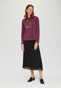 Bordeaux sweatshirt met capuchon en een patroon sjaal, gecombineerd met een geplooide zwarte midi rok. Glanzende zwarte loafers maken de outfit compleet.
