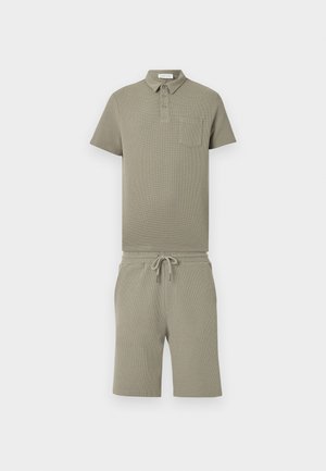 Kurzärmeliges olivgrünes Poloshirt mit Brusttasche, kombiniert mit passenden Shorts mit Kordelzug vor einfarbigem Hintergrund.