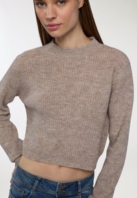 Młoda kobieta w beżowym, dzierganym swetrze na krótko i niebieskich dżinsach, pozująca z jedną ręką na biodrze na jednolitym tle.