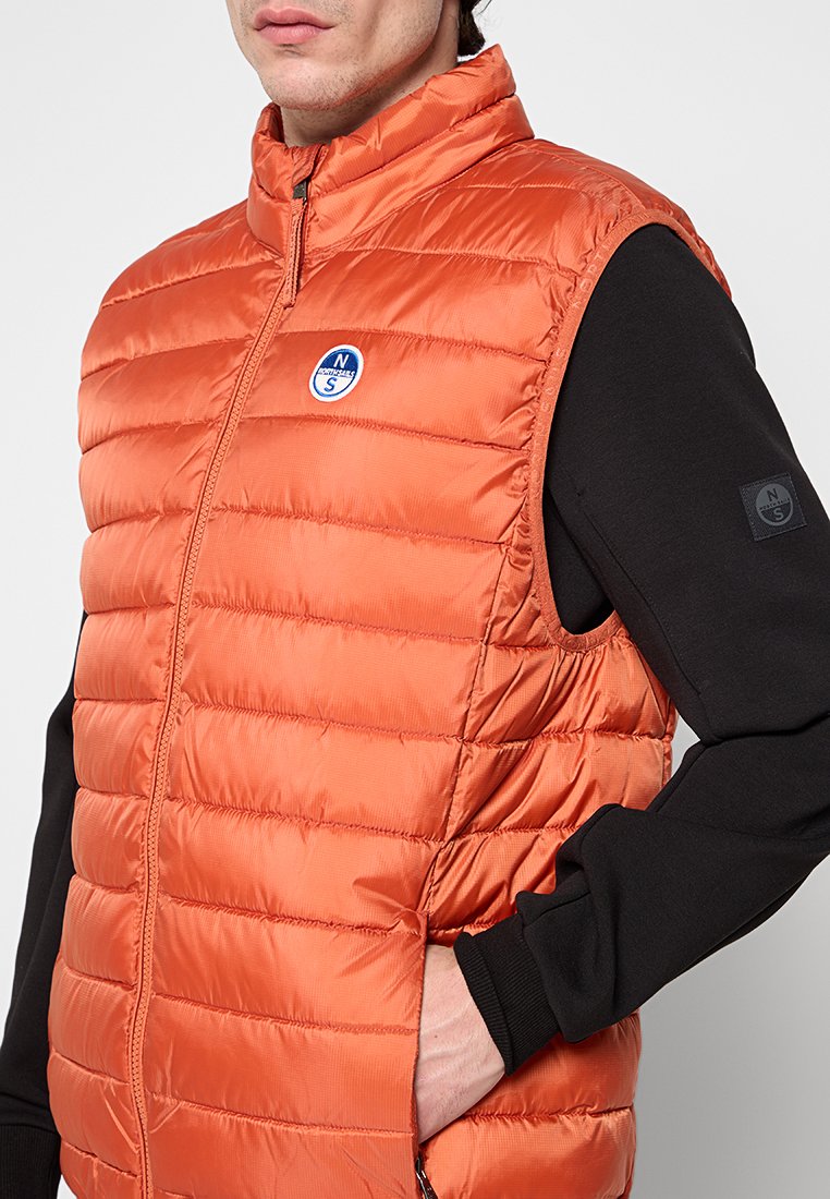 Orangene gesteppte Pufferweste mit hohem Kragen, Reißverschluss und rundem Logo. Schwarzes Langarmshirt darunter, sichtbare Tasche.