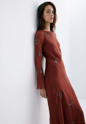 Femme portant une robe longue à manches longues couleur rouille avec broderies florales, appuyée contre un mur blanc, regardant vers la caméra.