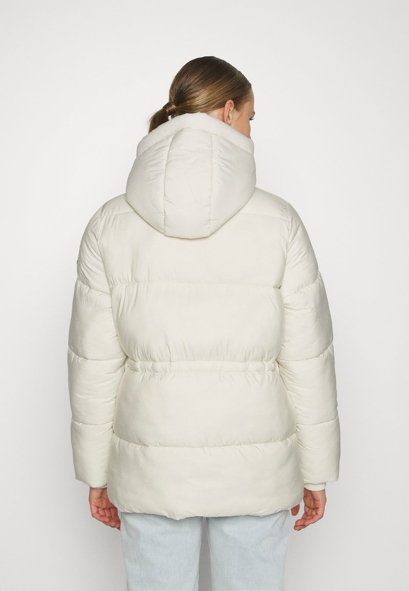 Chaqueta acolchada en crema claro, con capucha, textura acolchada y cintura ajustada para un fit entallado. Tela suave y lisa sin herrajes visibles.