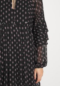 Robe noire transparente avec petits motifs floraux rouges et blancs, avec un col à nouer et des manches longues semi-transparentes à volants.