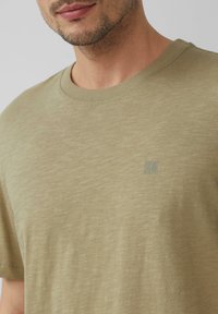 Oliwkowa bawełniana koszulka z krótkim rękawem i okrągłym dekoltem o gładkiej fakturze. Zawiera małe logo na lewej części klatki piersiowej.