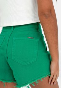 Pantaloni shorts in denim verde con orlo sfilacciato e una tasca posteriore. Presenta un piccolo etichetta del brand sulla tasca posteriore destra.