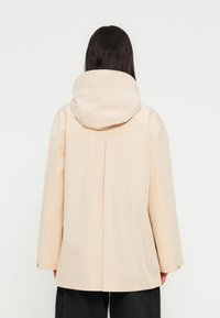 Veste de pluie beige avec une grande capuche, des manches larges et une ouverture à l'arrière. Matière lisse et légère avec des fermetures à pression aux poignets.