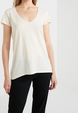 Frau trägt ein cremefarbenes T-Shirt mit kurzen Ärmeln und V-Ausschnitt und schwarze Hose, steht vor einem schlichten hellen Hintergrund.
