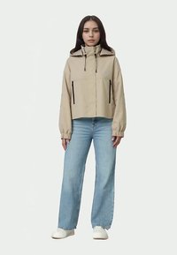 Noor noor seisab, seljas beež kapuutsiga jakk mustade tõmblukkudega, helesinised teksad ja valged kingad, lihtsa taustaga.